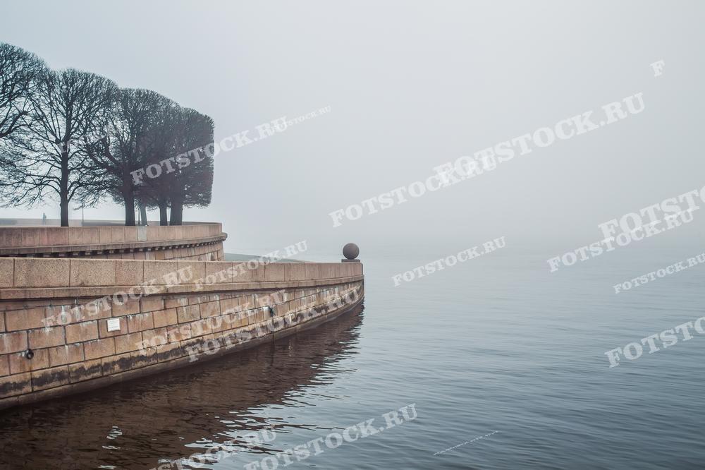 Фотография запечатлела набережную Васильевского острова в туманное утро, где тонкий слой тумана окутывает поверхность воды и расстилается вглубь кадра, сливаясь с небом. Каменные ступени набережной и гранитные шары ведут к группе деревьев, создавая впечатление уединенного и тихого уголка в городской среде. Сцена настолько спокойна, что вода кажется зеркальной. Такой снимок идеально подойдёт для использования в материалах о городском ландшафте, дизайне среды, а также в статьях о спокойствии и отдыхе, предлагая зрителям момент покоя и раздумий.